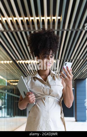 Angenehme, schwanger afroamerikanische Frau in kabellosen Ohrstöpseln mit lockigen Haaren, Textnachrichten auf dem Mobiltelefon in der Nähe des Glasgebäudes Stockfoto