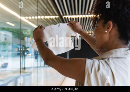 Rückansicht der Ernte lächelnde afroamerikanische Frau mit Babykleidung, die in der Nähe der Glaswand steht Stockfoto
