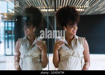 Angenehme, schwanger afroamerikanische Frau in kabellosen Ohrstöpseln mit lockigen Haaren, Textnachrichten auf dem Mobiltelefon in der Nähe des Glasgebäudes Stockfoto