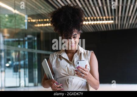 Angenehme, schwanger afroamerikanische Frau mit lockigen Haaren, Textnachrichten auf dem Mobiltelefon, während sie ihren Laptop in der Nähe des Glasgebäudes trug Stockfoto