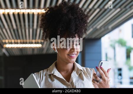 Angenehme, schwanger afroamerikanische Frau in kabellosen Ohrstöpseln mit lockigen Haaren, Textnachrichten auf dem Mobiltelefon in der Nähe des Glasgebäudes Stockfoto