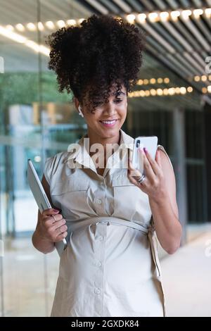 Angenehme, schwanger afroamerikanische Frau in kabellosen Ohrstöpseln mit lockigen Haaren, Textnachrichten auf dem Mobiltelefon in der Nähe des Glasgebäudes Stockfoto