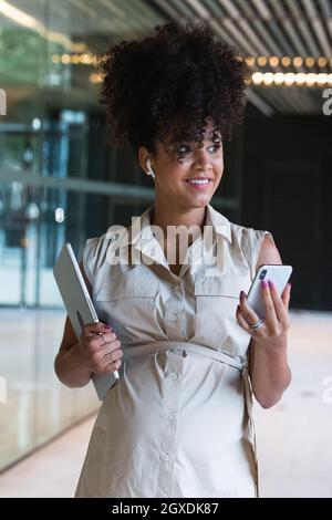 Angenehme, schwanger afroamerikanische Frau in kabellosen Ohrstöpseln mit lockigen Haaren, Textnachrichten auf dem Mobiltelefon in der Nähe des Glasgebäudes Stockfoto