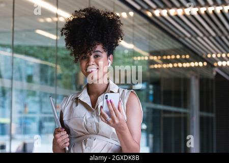 Angenehme, schwanger afroamerikanische Frau in kabellosen Ohrstöpseln mit lockigen Haaren, Textnachrichten auf dem Mobiltelefon in der Nähe des Glasgebäudes Stockfoto