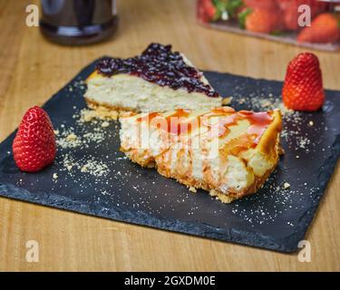 Set von Käsekuchen mit Erdbeer, Erdbeersoße, zerbröckeltem Gebäck und zwei Erdbeeren auf einem schwarzen Schieferbrett. Stockfoto