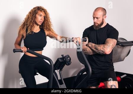 Konzentrierte Sportlerin mit langen lockigen Haaren, die auf dem Fahrradcomputer sitzt und mit einem Personal Trainer im Fitnessstudio Cardio-Training macht Stockfoto