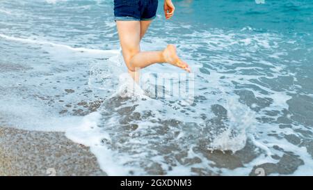Barfuß beschnittene, nicht erkennbare weibliche Reisende, die bei windigem Wetter am Sandstrand entlang läuft, der von schaumigen Wellen gewaschen wird Stockfoto