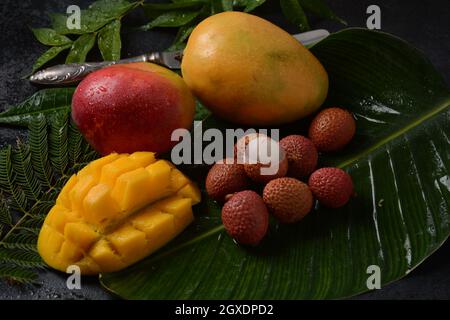 Frische Mangofrucht mit in Scheiben geschnittenen Mangostückchen auf einem Teller und frischer und geschälter Litschi, die die rote Haut und das weiße Fleisch auf dunklem Hintergrund zeigt. Stockfoto