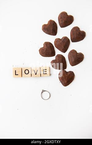 Trüffelschokolade in Form eines Herzens auf weißem Hintergrund. Inschrift Love aus Holzbuchstaben und einem Ring mit einem Diamanten. Hand und Hand Stockfoto