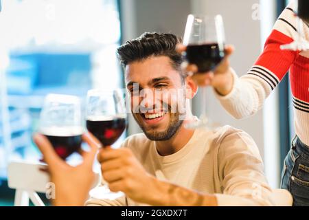 Fröhlicher junger Mann, der mit Freunden mit einem Glas Rotwein toasten feiert, während sie ihre Weingläser in Anrede mit Fokus auf sein Gesicht heben Stockfoto