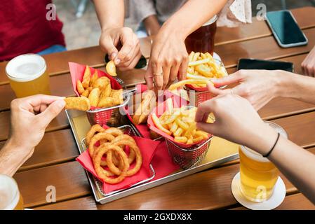 Ein hoher Winkel der Erntegut unkenntliche Freunde, die Pommes Frites und knusprige Zwiebelringe vom Tablett nehmen, während sie sich zusammentun und Bier trinken Stockfoto