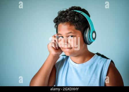 Fröhlicher, intelligenter, lockiger afroamerikanischer Junge mit jungen Haaren, der über blaue kabellose Kopfhörer Lieblingsmusik genießt und im Studio auf die Kamera schaut Stockfoto