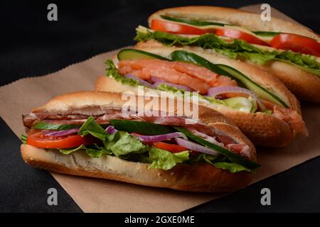 Frische U-Boot-Sandwiches mit Speck, Käse, Tomaten, Salat, Gurken und Zwiebeln auf dunklem Hintergrund Stockfoto