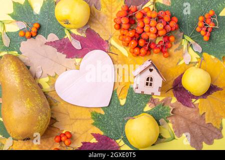 Holzherz, die Form eines Holzhauses auf mehrfarbigen roten, orangefarbenen, grünen, trocken gefallenen Herbstblättern und orangen Vogelbeeren, Äpfeln und Birnen auf einem Stockfoto