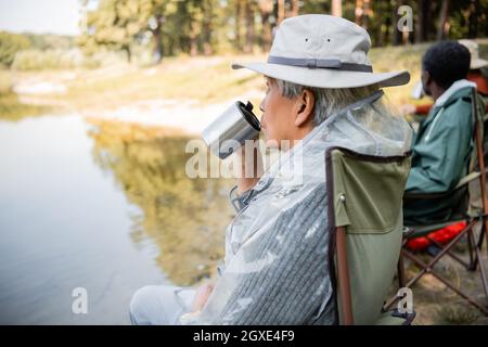 Senior Asian Mann hält Thermobecher in der Nähe verschwommener Freunde in Angel-Outfit in der Nähe von See Stockfoto