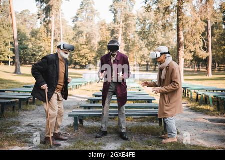 Aufgeregt Senior Mann mit Gehstock mit vr-Headset in der Nähe von interracial Freunde im Park Stockfoto
