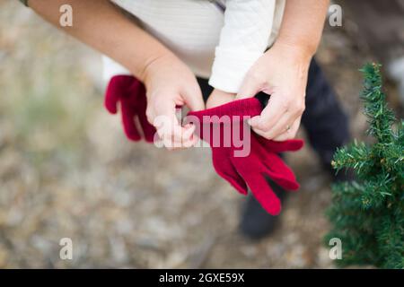 Fürsorgliche Mutter Kind in der Nähe von kleinen Weihnachtsbaum abstrakt rote Handschuhe anziehen. Stockfoto