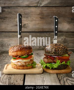 Zwei Cheeseburger mit Gemüse und Fleischsteak auf einem Holzbrett, ein Sandwich wird mit einem Messer durchbohrt Stockfoto