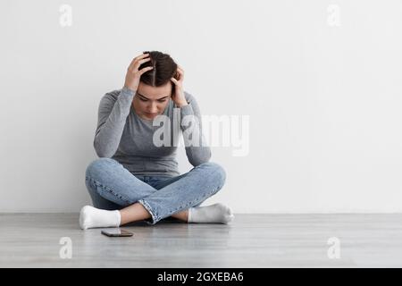 Verzweifelte, unglückliche, traurige kaukasische, tausendjährige weibliche Frau, die an Depressionen litt und den Kopf mit der Hand hielt und auf dem Boden saß Stockfoto