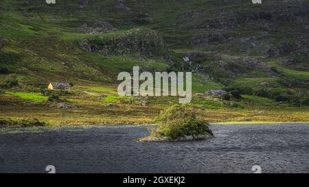 Kleine felsige Insel mit verdrehten Bäumen am Lough Gummeenduff mit Häusern auf dem Hügel von MacGillycuddys Reeks Mountains, Ring of Kerry, Irland Stockfoto