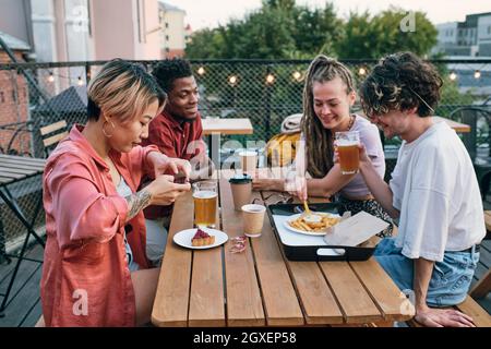 Glückliche junge interkulturelle Freunde, die im Café im Freien Fast Food und Getränke am Tisch genießen Stockfoto