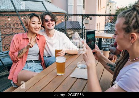 Glückliches junges interkulturelles Paar posiert für ihren Freund mit dem Smartphone, während es sich im Café im Freien entspannt Stockfoto