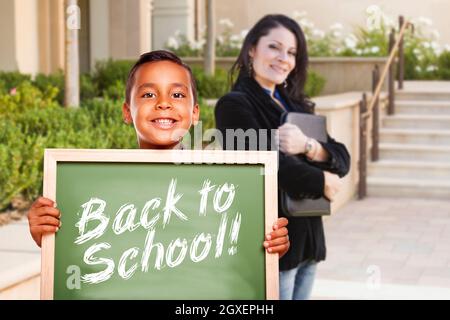 Glücklicher hispanischer Junge, der draußen auf dem Schulgelände die Kreidetafel der Schule zurückhält, während der Lehrer darauf schaut. Stockfoto