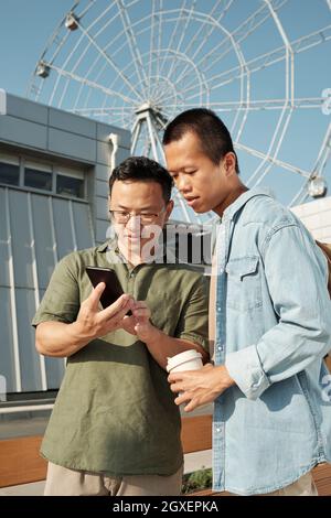 Zwei chinesische Geschäftsleute in Casualwear, die den Smartphone-Bildschirm gegen den Vergnügungspark betrachten Stockfoto