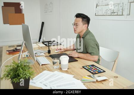 Geschäftige junge kreative Designerin, die im Büro vor einem Computermonitor sitzt Stockfoto