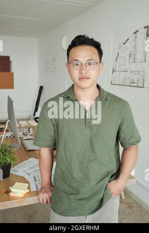 Der junge Profi-Designer steht mit einem Computermonitor gegen seinen Arbeitsplatz Stockfoto