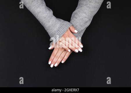 Weibliche Hände in grauem Strickpullover mit schöner Maniküre - weiße Nägel auf schwarzem Hintergrund, Banner mit Kopierfläche. Nagelpflegekonzept Stockfoto