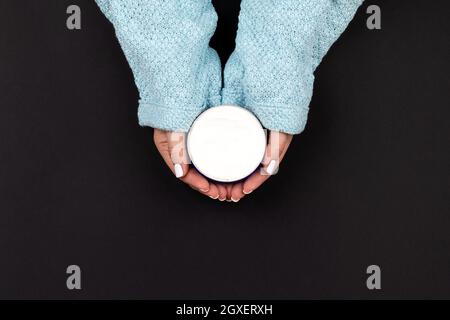 Weibliche Hände in blau gestrickt Pullover mit weißen Nägeln Maniküre halten Glas mit Creme auf schwarzem Hintergrund. Hautpflege, Bio-Kosmetik-Konzept, top vi Stockfoto