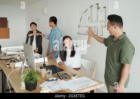 Glückliche junge Kollegen, die vor dem Hintergrund von zwei anderen Kollegen eine Geste mit fünf hochgestümmelt haben Stockfoto