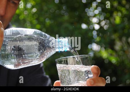 Wasser in ein Glas in der Hand mit Garten im Hintergrund gegossen. Erfrischung mit sauberem, natürlichem Mineralwasser aus einer Plastikflasche. Stockfoto