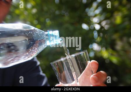 Wasser in ein Glas in der Hand mit Garten im Hintergrund gegossen. Erfrischung mit sauberem, natürlichem Mineralwasser aus einer Plastikflasche. Stockfoto