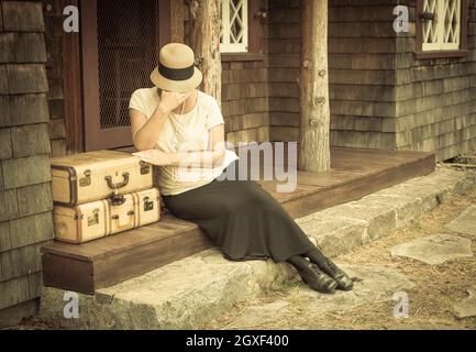 Distressed 1920er Jahre gekleideten Mädchen neben Koffer auf Veranda mit Vintage-Effekt hinzugefügt. Stockfoto