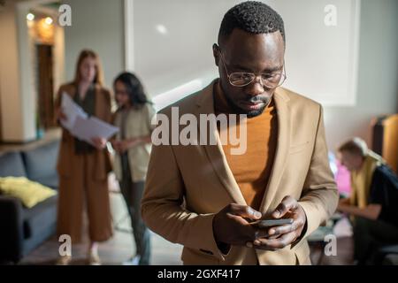 Serious African Geschäftsmann scrollt im Smartphone gegen Gruppe von kaukasischen Kollegen Stockfoto