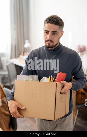 Junger Geschäftsmann, der einen Karton mit Vorräten trägt, während er seinen Arbeitsplatz wechselt oder sein Büro verlässt Stockfoto