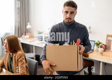 Junger unzufriedener Mitarbeiter hält einen Karton mit Büromaterial, während er vor der Kamera steht Stockfoto