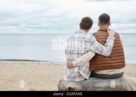 Rückansicht Portrait eines jungen schwulen Paares, das sich am Strand umarmt und dabei Meerblick und Platz zum Kopieren genießt Stockfoto