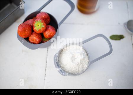 Frische Erdbeeren und Mehl in Messbechern neben Heidelbeeren und Honig. Gesunde Zutaten in Behältern über dem weißen Zähler. Gesunde Mahlzeit pro Stockfoto