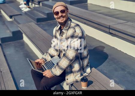 Lächelnder junger Mann blickt auf die Kamera, hält den Laptop in den Händen und sitzt draußen am Strand Stockfoto