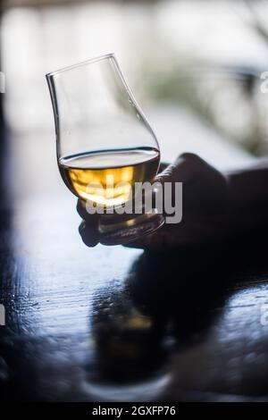 Nahaufnahme einer Hand, die ein Glencairn Whisky-Glas auf einem Holztisch hält, mit geringer Schärfentiefe. Stockfoto