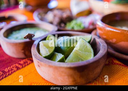 Nahaufnahme von Tonschüsseln mit Limettenscheiben, grüner Sauce und Lammbrühe mit verschwommenem, gehacktem Fleisch als Hintergrund. Gewürzen und geschnittenes Gemüse über oran Stockfoto