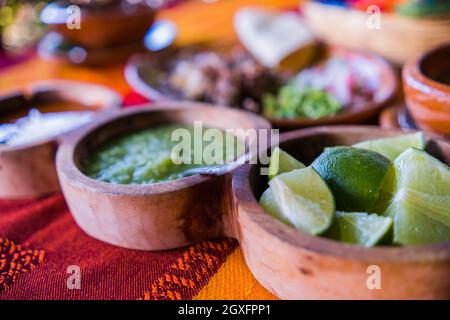 Nahaufnahme von Tonschüsseln mit Limettenscheiben und heißen grünen und roten Saucen mit verschwommenem, gehacktem Fleisch als Hintergrund. Gewürzen und geschnittenes Gemüse über oran Stockfoto
