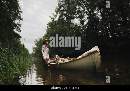 Der junge Mann steht im seichten Fluss neben seinem Kanu mit einem Hund Stockfoto