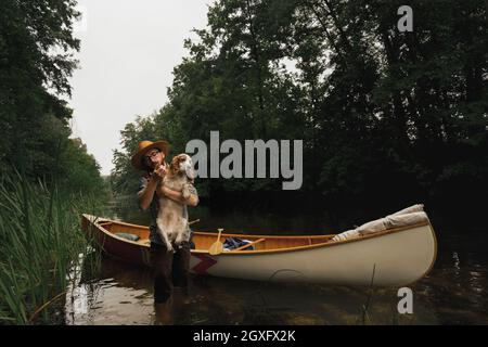 Der junge Mann steht im seichten Fluss neben seinem Kanu mit einem Hund. Abenteuerliches Konzept für aktive Erholung Stockfoto