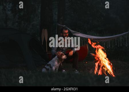 Person sitzt neben seinem Hund am Lagerfeuer, Camping mit Kanu in schöner Natur Stockfoto