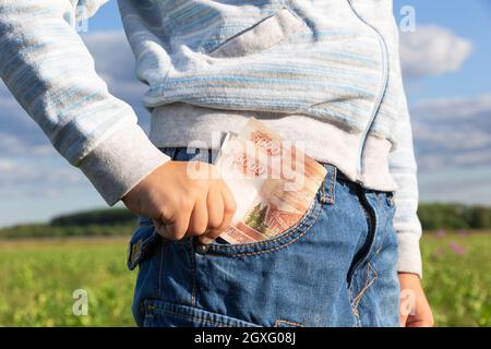 Die Hand eines Kindes nimmt an einem hellen sonnigen Sommertag Papiergeld-Rubel aus der Jeanentasche auf dem Hintergrund des blauen Himmels. Selektiver Fokus. Nahaufnahme Stockfoto