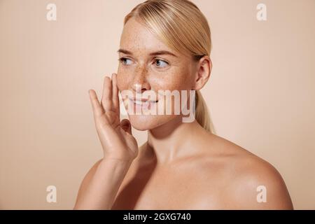 Lächelnde Frau berührt ihr Gesicht mit dem Finger, während sie im Studio vor einem pastellfarbenen Hintergrund steht Stockfoto